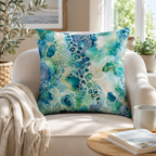 An abstract emerald green and gold decorative throw pillow resting on a modern cream-colored accent armchair, with a cozy knit blanket draped over the side and a sunlit window in the background.