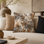 A close-up of an abstract wild floral and leopard-spotted accent pillow arranged on a neutral linen sofa, paired with brown and black textured cushions in a warm minimalist interior.