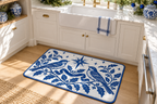 Blue & White Christmas Songbird Printed Floor Mat floor mat, lifestyle view 1: A bright American kitchen with white shaker cabinets, pale oak floor and blue-and-white ceramics on an open shelf, the reference small landscape floor mat laid flat before the sink, all four rounded corners visible.