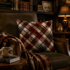 A classic tartan plaid accent pillow arranged on a vintage dark leather armchair in a cozy dimly lit library, with a classic desk lamp and bookshelf in the background.