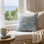 A coastal ocean-inspired throw pillow placed on a sunlit white window seat overlooking the blue sea, paired with a ceramic coffee mug, books, and a soft throw blanket.
