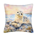 A square ocean-themed pillow cover isolated on a white background, featuring a vivid watercolor painting of a cute harbor seal resting on coastal rocks under a pastel pink and yellow sunset sky.