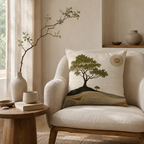 A zen-inspired scenic throw pillow with an artistic tree and warm sun design, arranged on a cozy beige armchair next to a wooden side table with a simple bud vase.