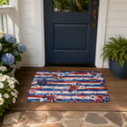 An Americana independence day themed doormat laid out on a dark wooden porch in front of a navy blue front door, accented by white daisies, blue hydrangeas, and a potted green fern.