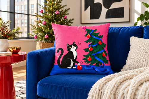 Christmas Cat & Tree Printed Pillow Cover pillow covers, lifestyle view 1: A colorful contemporary American apartment living room with a cobalt-blue sofa and one exact reference cat pillow clearly displayed against its back, a bright red side table and an abstract black-and-white wall print.