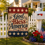 Patriotic seasonal garden flag with elegant cursive "God Bless America" text, placed by a traditional white mailbox, white picket fence, and blooming red and blue flowers.